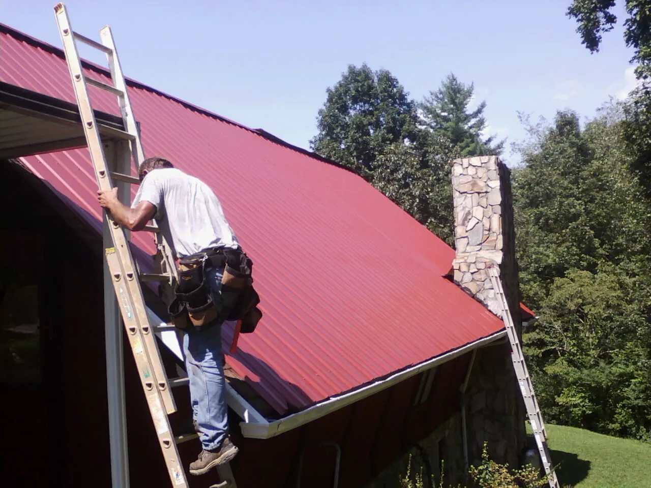 Roofer on a ladder beside a red metal roof and stone chimney — Soffit & Fascia Repair by Roofing Brothers in Saint Martinville